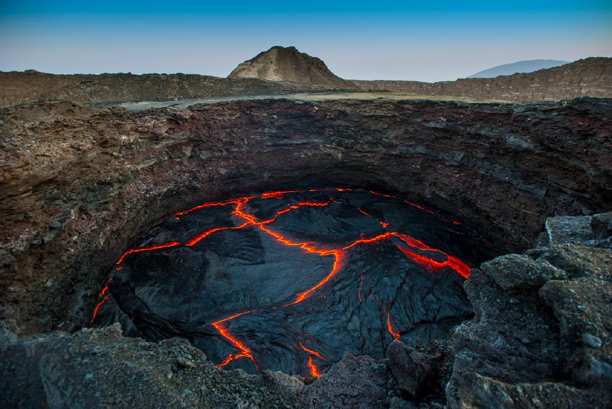 A high-angle landscape shot looking into a volcanic crater filled with active, glowing red lava, surrounded by dark, rugged, rocky terrain under a clear blue sky