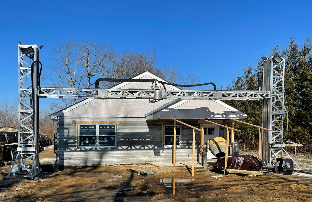 A large-scale 3D concrete printer constructing a residential home on a bright sunny day with a blue sky background.