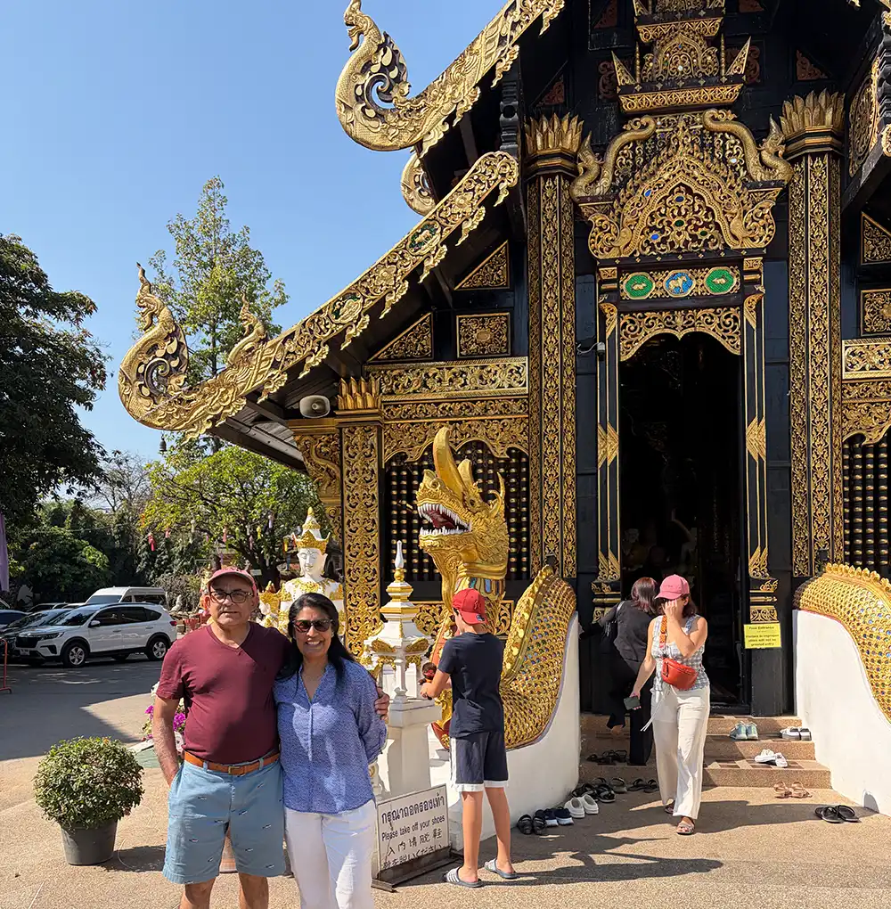 A full-shot photograph of Chandu and Hima Patel standing and smiling together in front of an ornate, black and gold Thai temple in Chiang Mai, Thailand. The temple features intricate gold carvings, a tiered roof with decorative finials, and a large golden naga statue guarding the entrance.