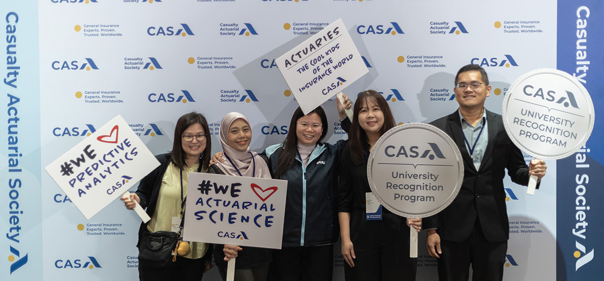 Five Casualty Actuarial Society (CAS) University Recognition Program members holding playful promotional signs in front of a CAS logo signage backdrop